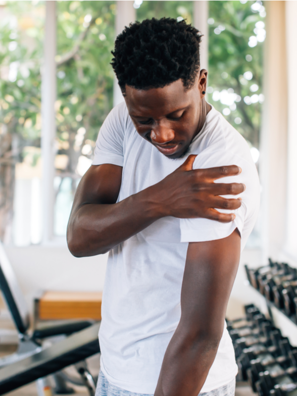 Side view of muscular African American man standing and suffering from shoulder pain during workout with dumbbells.